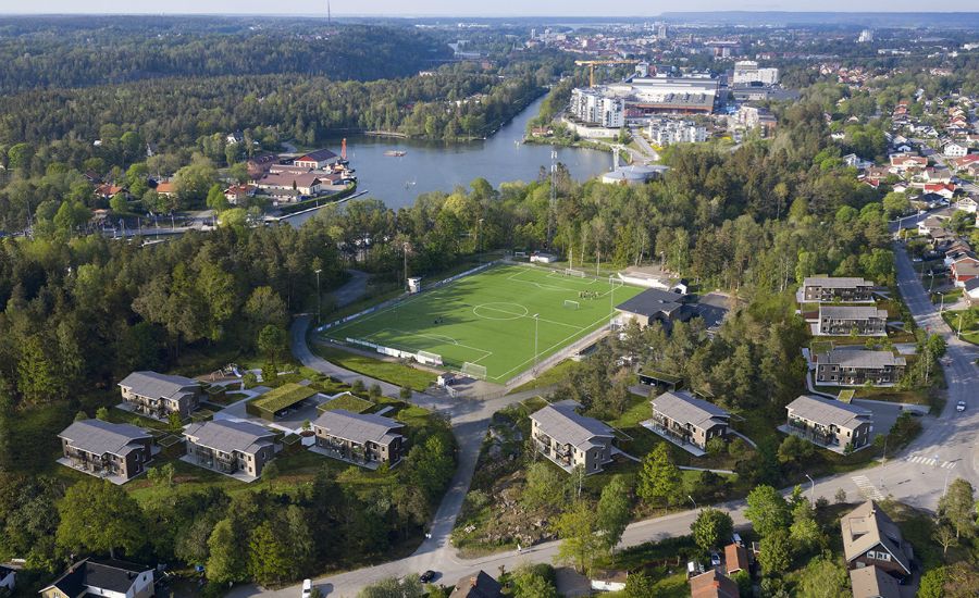 Luftvy av ett grönskande område med fotbollsplan, bostadshus, skog och sjö i bakgrunden