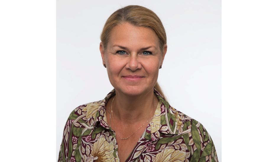 Kvinna med blont hår, iförd en skjorta med färgglatt blommönster, står mot en vit bakgrund