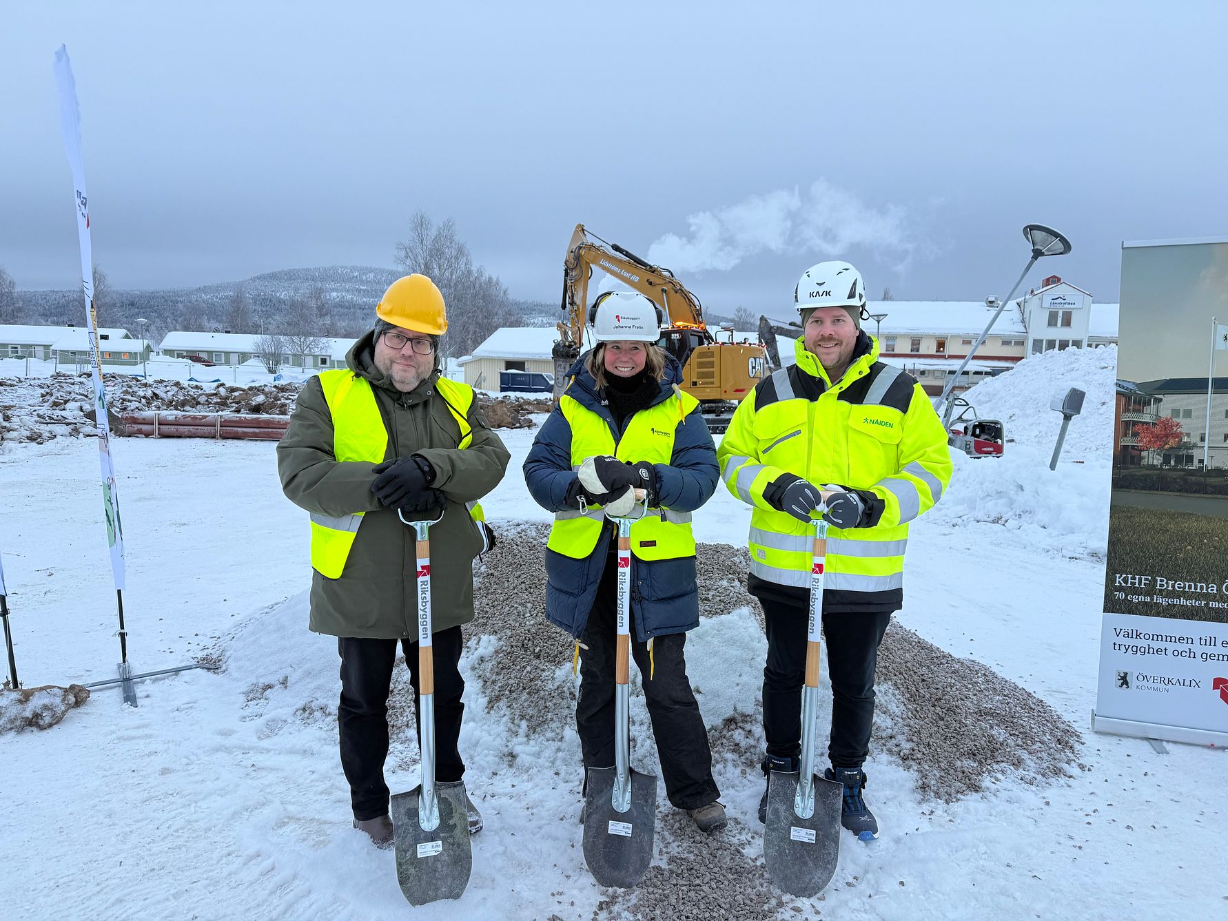 Tre personer i varselkläder och hjälmar håller i spadar vid en byggarbetsplats med snötäckt mark och grävmaskin i bakgrunden