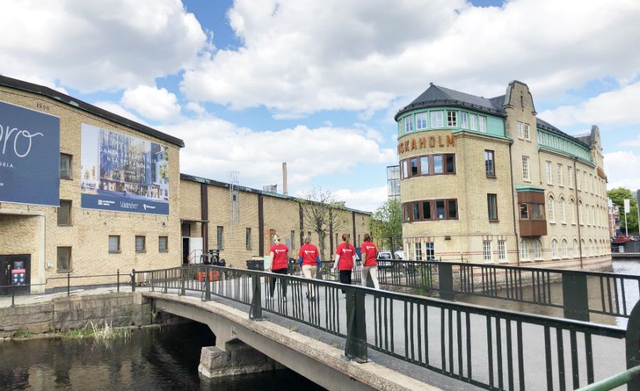 Bro som sträcker sig över en vattenkanal med tegelbyggnader och skyltar i bakgrunden, samt människor i röda tröjor som går på bron