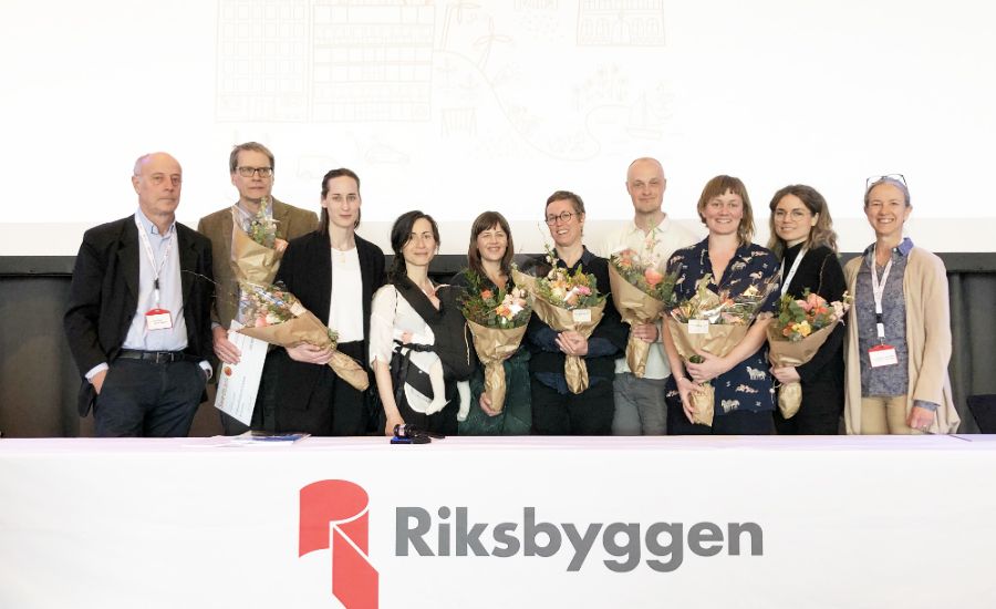 En grupp personer står framför ett bord med texten "Riksbyggen" och håller buketter med blommor, med en ljus bakgrund