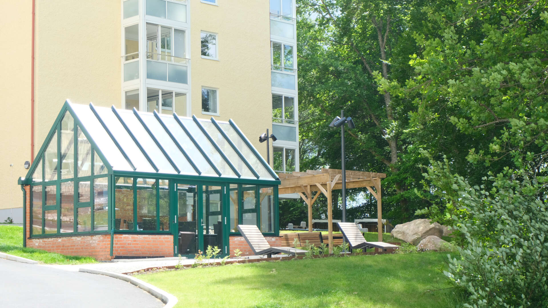 Växthus och pergola med solstolar framför ett flerbostadshus i en grönskande miljö