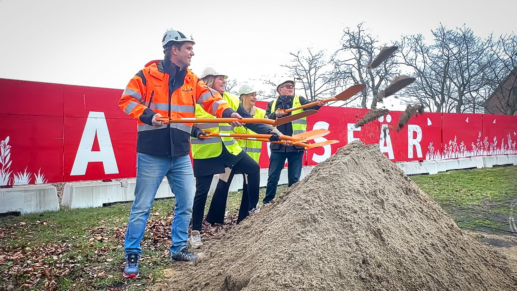 Fyra personer i reflexvästar och hjälmar som spadar sand vid en byggarbetsplats