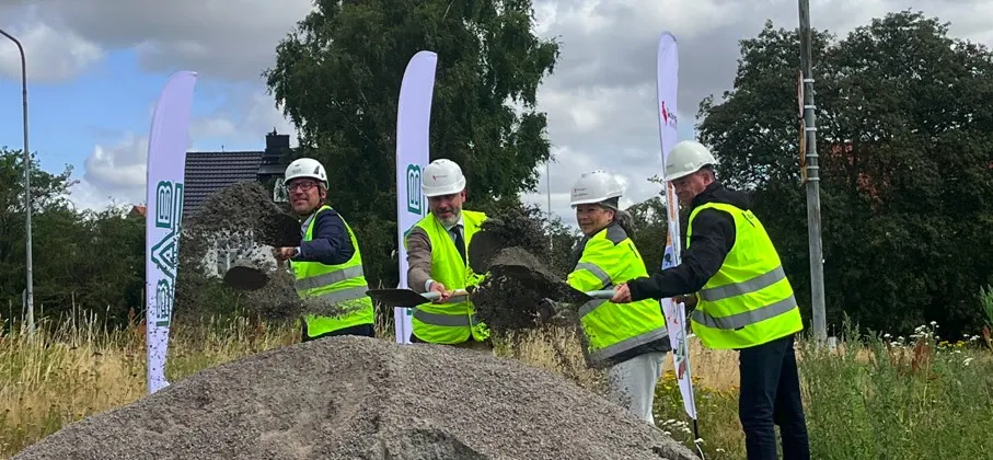 Fyra personer i reflexvästar och bygghjälmar skyfflar jord vid en byggplats med Bostadsrättsföreningen Riksbyggen-flaggor