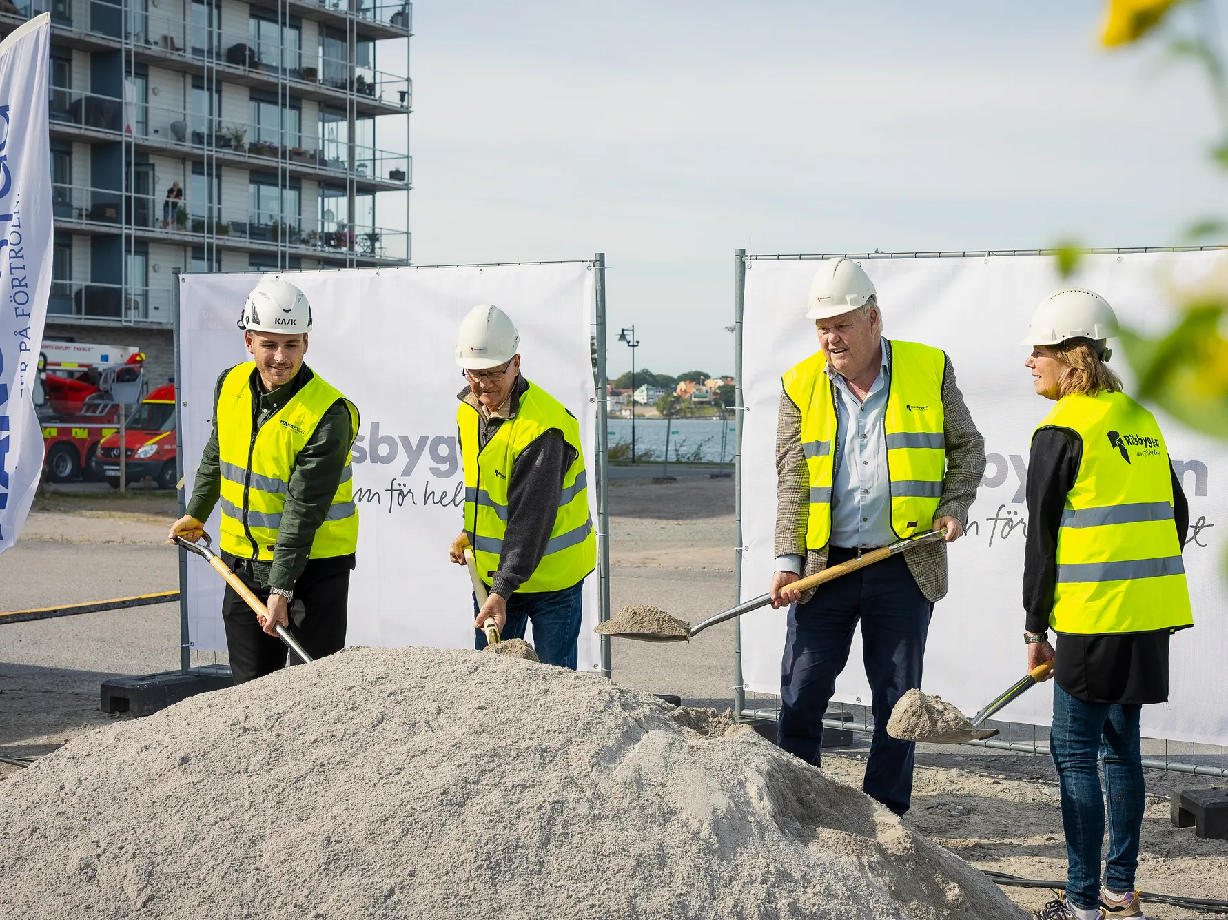 Fyra personer i hjälm och reflexväst skyfflar sand vid ett byggprojekt