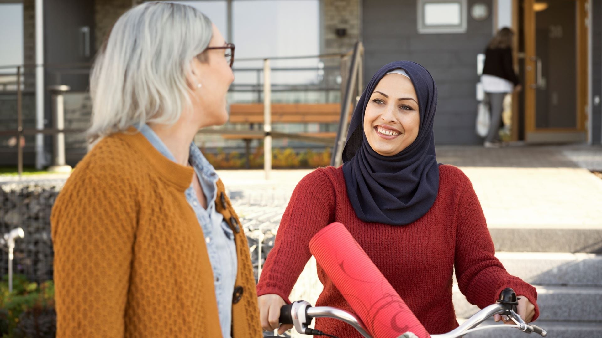 Två kvinnor pratar och ler utanför en byggnad, en av dem håller i en cykel med en röd yogamatta