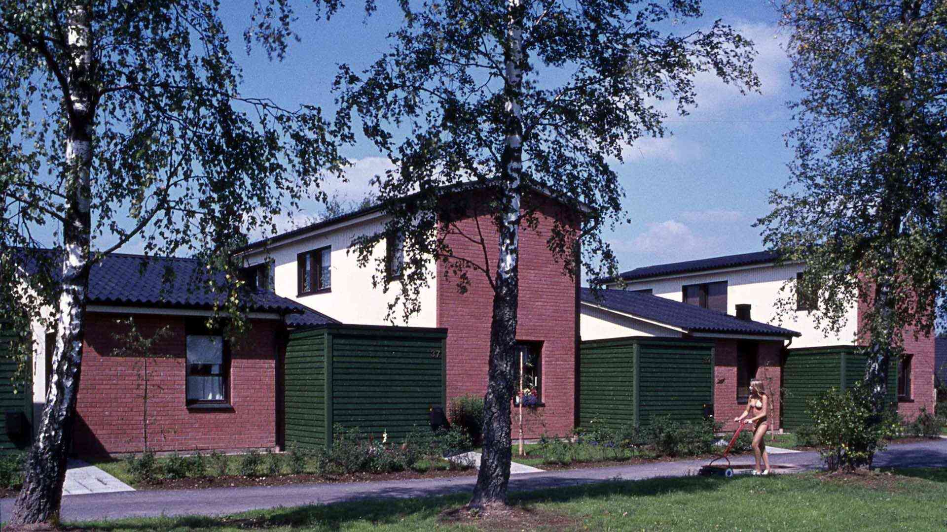 Radhus med röda tegelväggar och gröna staket, träd i förgrunden, en person klipper gräset med en handgräsklippare