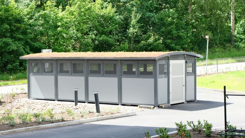 Grå byggnad med grönt tak i en grönskande omgivning, omgiven av asfalt och växtlighet