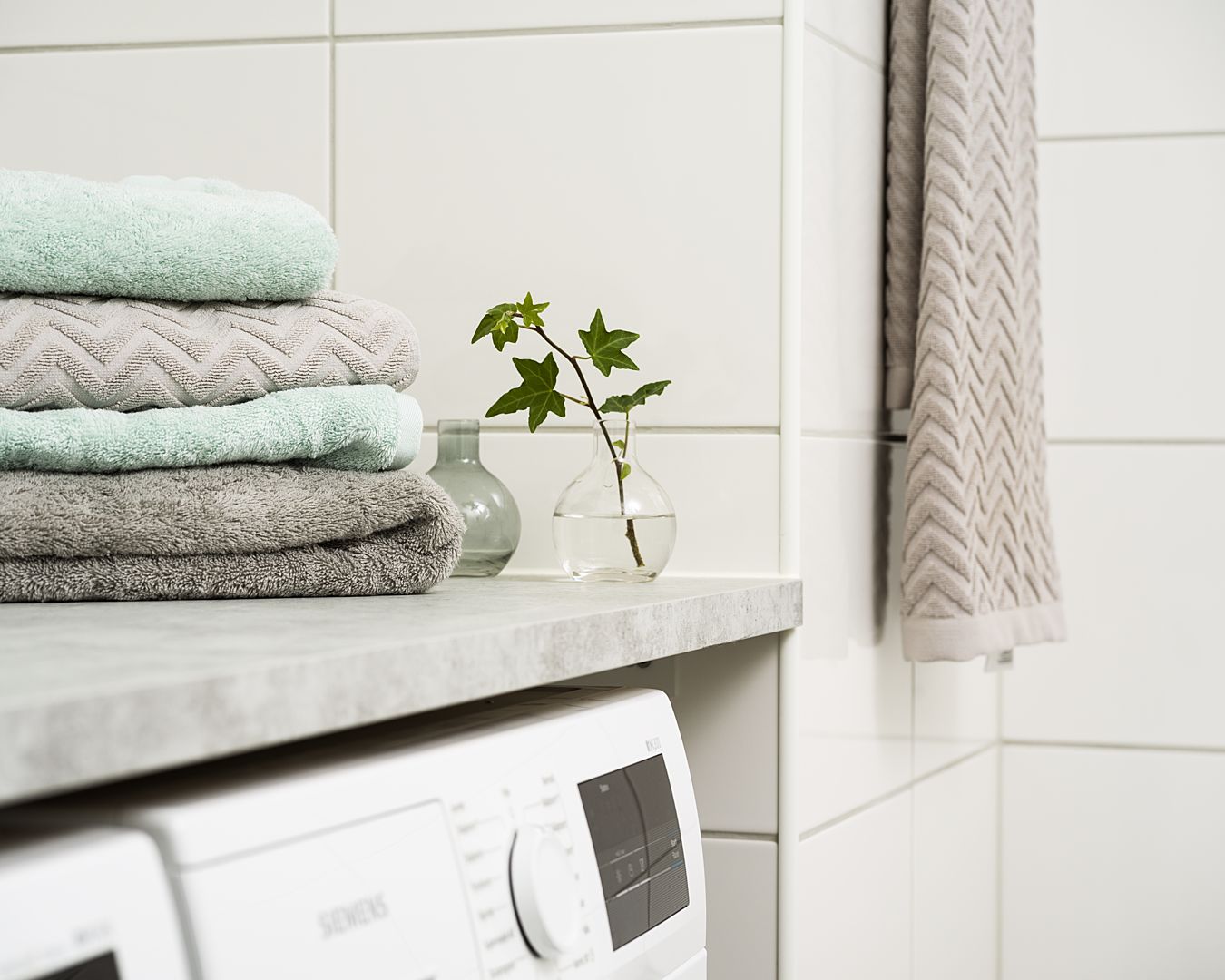 Vita och grå handdukar staplade på en bänkskiva bredvid en liten vas med gröna växter, framför en tvättmaskin i ett kaklat badrum