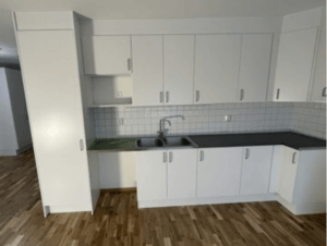 Vit köksinredning med flera skåp, diskho, grå bänkskivor och kaklat stänkskydd. Golvet är av trä och rummet har naturligt ljus