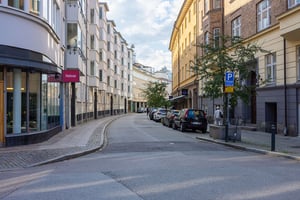 Gata med parkerade bilar på båda sidor, omgiven av flerbostadshus och butiker under en klar himmel