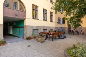 Innergård med stenlagd mark, sittgrupp med träbord och bänkar, blomkrukor, cyklar och grönska framför en gul byggnad med fönster