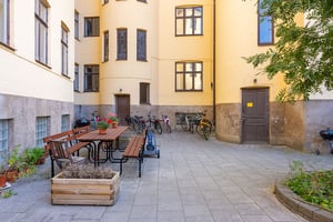 Innergård med cyklar, bänkar, blommor och en planteringslåda framför en gul byggnad med bruna fönsterkarmar