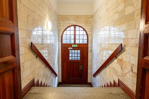 Trappa med vita kaklade väggar, träräcken och en mörkbrun dörr med glasrutor vid slutet av trapphuset