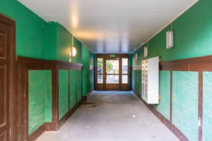 Grönmålad korridor med bruna trädetaljer, postboxar på ena väggen och glasdörrar i bakgrunden som leder ut till en gård