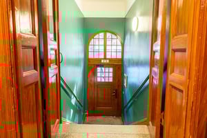 Trappa med träpaneler och gröna väggar, leder ner till en dörr med välvt fönster och glasruta. Ljuskälla synlig på väggen