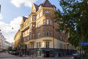 Gulbrunt flerfamiljshus i tegel med spetsiga tak, stora fönster och butiker på bottenvåningen, beläget vid en gatukorsning med omgivande grönska och parkeringsskylt