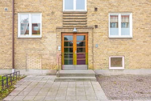 Entré till en byggnad med ytterdörr i trä, omgiven av gula tegelväggar, fönster och en stenbelagd gång