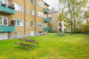 Bostadshus med tegelfasad och gröna balkonger, omgivna av en gräsmatta med picknickbord och träd