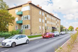 Flerfamiljshus i tegel med balkonger, parkerade bilar längs en asfalterad gata och grönska i förgrunden