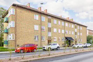 Brun tegelbyggnad med flera våningar, fönster och balkonger, bilar parkerade framför på en asfalterad väg