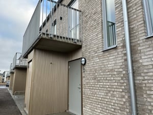 Modern byggnad med fasad av ljusa tegelstenar, balkong med metallräcken och en dörr med en rund utomhuslampa bredvid