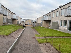 Gångväg mellan radhus med gräsmattor, nyplanterade buskar och betongfasader under en molnig himmel