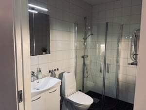 Badrum med vitt kakel, dusch med glasväggar, toalett och handfat med spegel ovanför