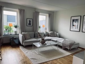 Ljust vardagsrum med hörnsoffa, soffbord i glas och väggdekorationer. Rummet har stora fönster, gardiner och en ljus matta på parkettgolv