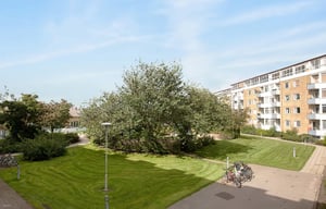 Utemiljö med grönskande träd, gräsmattor, gångvägar och cykelställ framför flerfamiljshus under en klar blå himmel