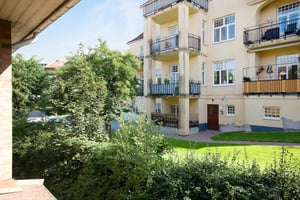 Gul lägenhetsbyggnad med balkonger, omgiven av grönska och en gräsmatta under en klar himmel