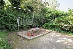 Sandlåda med leksaker som spadar och hinkar, omgiven av grönska, med en bänk och en klätterställning i bakgrunden