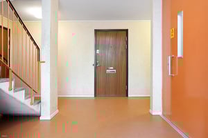 Entréområde med en trappa till vänster, en brun dörr i mitten och en orange dörr till höger. Väggarna är ljusa och golvet är i orange ton