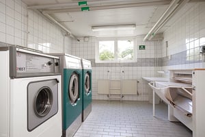 Tvättstuga med vitkaklade väggar, tre tvättmaskiner, ett torkskåp och ett fönster som släpper in naturligt ljus