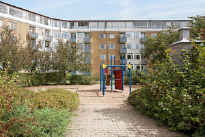 Lekplats med sand och lekutrustning omgiven av buskar, träd och ett flerbostadshus i bakgrunden