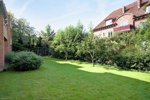 Grön gräsmatta omgiven av buskar och träd, med bostadshus i bakgrunden