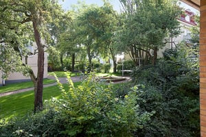 Grönskande trädgård med träd, buskar och gräsmatta omgiven av byggnader. En gångstig leder genom området