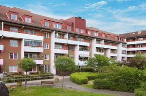 Flerfamiljshus med röda tak och vit-röda fasader, omgivna av grönskande trädgård och gångvägar