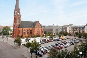 Röd tegelkyrka med högt torn och klocka, omgiven av parkerade bilar och tält på ett torg i stadsmiljö