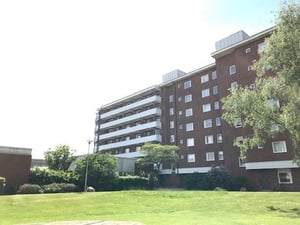 Brun flerfamiljsbyggnad med flera våningar, omgiven av grönskande gräsmatta och träd under en klar himmel