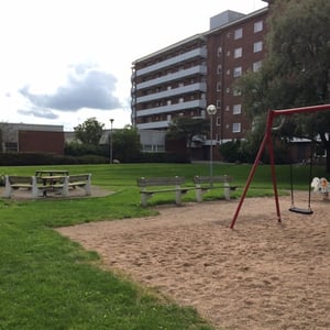 Lekplats med gungställning, sandunderlag och träbänkar, omgiven av gräsmatta. En flerfamiljsbyggnad syns i bakgrunden
