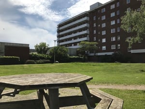 Träbord på en grön gräsmatta framför ett flerfamiljshus med tegelväggar under en blå himmel med moln