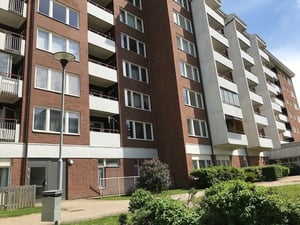 Flerbostadshus i tegel och vit fasad med balkonger, omgiven av grönska och en gångväg