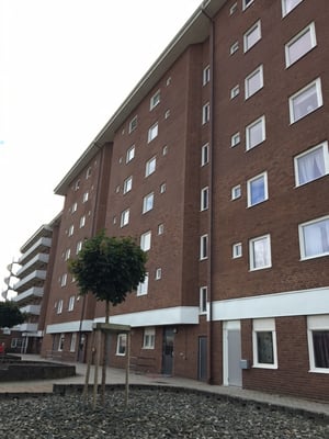 Brun tegelbyggnad med flera våningar och vita fönster, omgiven av små träd och stenbelagd mark