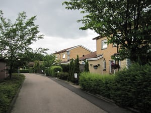 Rad av tegelhus med trädgårdar, gröna buskar och träd längs en asfalterad gångväg under molnig himmel