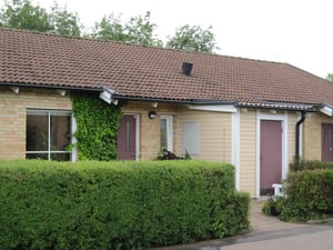 En enfamiljsvilla med tegelväggar och ett rött tegeltak, omgiven av en grön häck och träd i bakgrunden