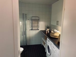 Badrum med duschkabin, svart golv, vita väggplattor, tvättmaskin, torktumlare och hyllor med korgar