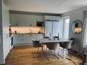 Modernt kök med vita skåpluckor, rostfria vitvaror och en matplats med ett bord och sex stolar. Rummet har trägolv, ett stort fönster och en rund spegel på väggen