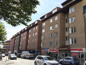 Flerbostadshus i tegel bredvid en gata med bilar, butiker och träd i förgrunden