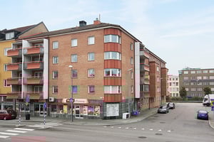 Fyravånings bostadshus i tegel med balkonger, butik på bottenvåningen och parkerade bilar längs gatan